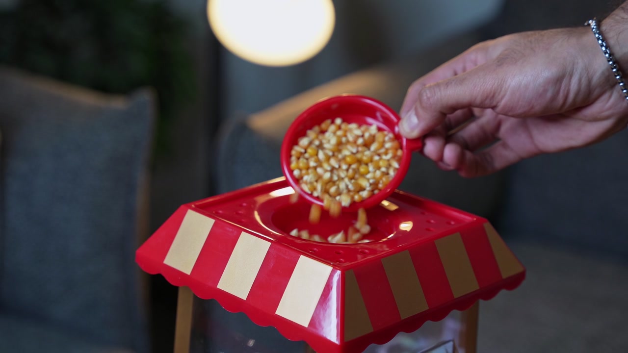 une main versant des grains de maïs séchés d'une tasse à mesurer rouge dans une machine à popcorn rétro. Éclairage intérieur chaleureux à la maison lors de la préparation d'une soirée cinéma dans un salon moderne