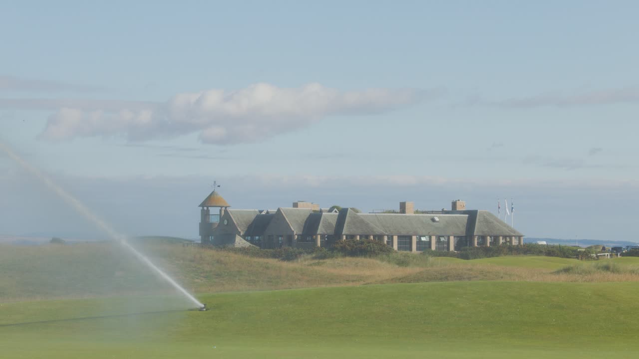 A wide shot shows a golf course green being watered by a rotating sprinkler under daylight, with a large historic clubhouse and blue sky in the background