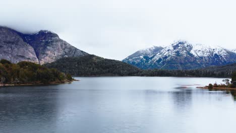 From-the-sky,-a-serene-lake-accompanied-by-the-stunning-beauty-of-the-snow-capped-peaks-and-lush-forested-slopes-of-Patagonia