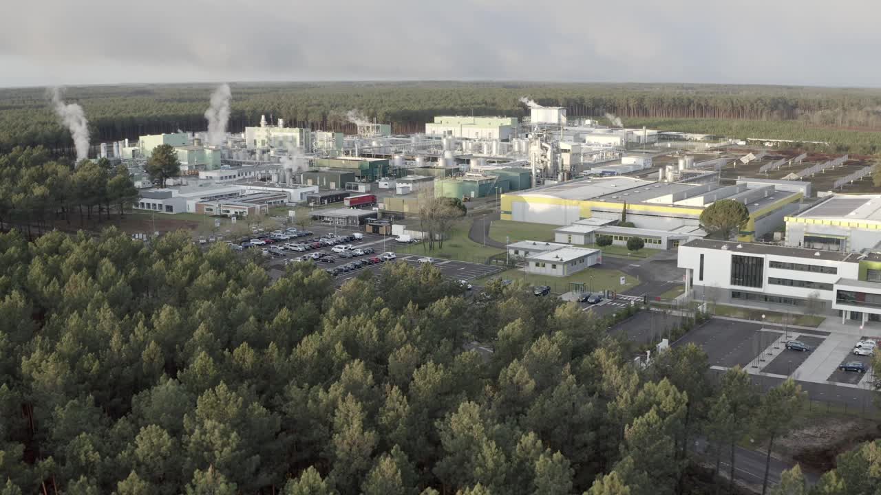 Drone establishing shot of large industrial plant with parking area, bordering pine forest, steam visible, Castets, France