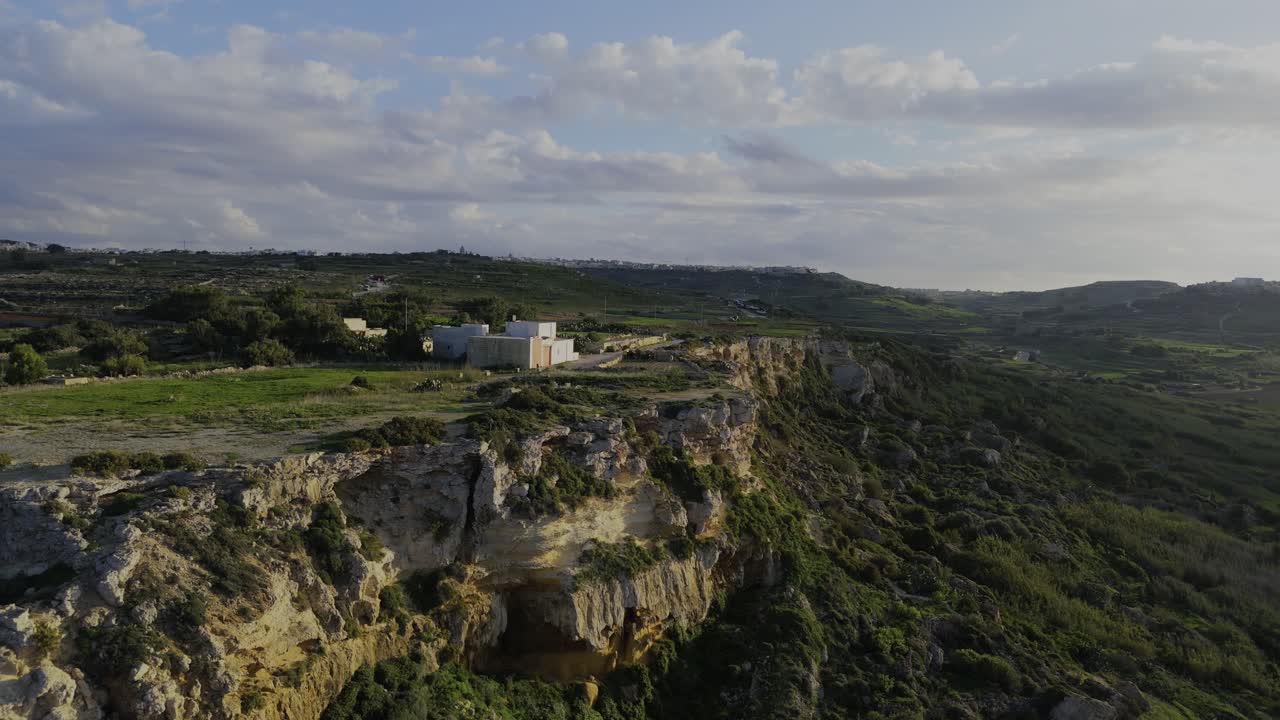 A dynamic drone move flying left and backward while turning right reveals the Mixta Cave cliffs, shifting toward a warm sunset view over Ramla Beach on Gozo