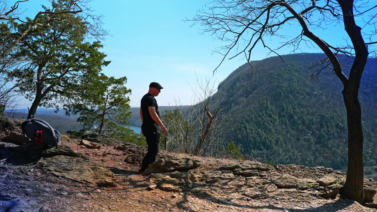 New Jersey, USA, 1 maggio 2026: Vista posteriore di un escursionista che riposa su una cima rocciosa di montagna. Escursionista con attrezzatura seduto su una sporgenza di pietra che si affaccia su un canyon fluviale e colline boscose sotto un cielo azzurro e limpido.