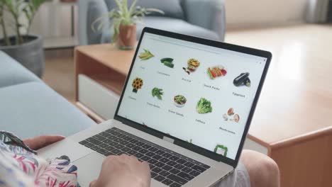 asian man sitting on couch and using laptop computer buys in internet shop. male selects vegetables in grocery online store