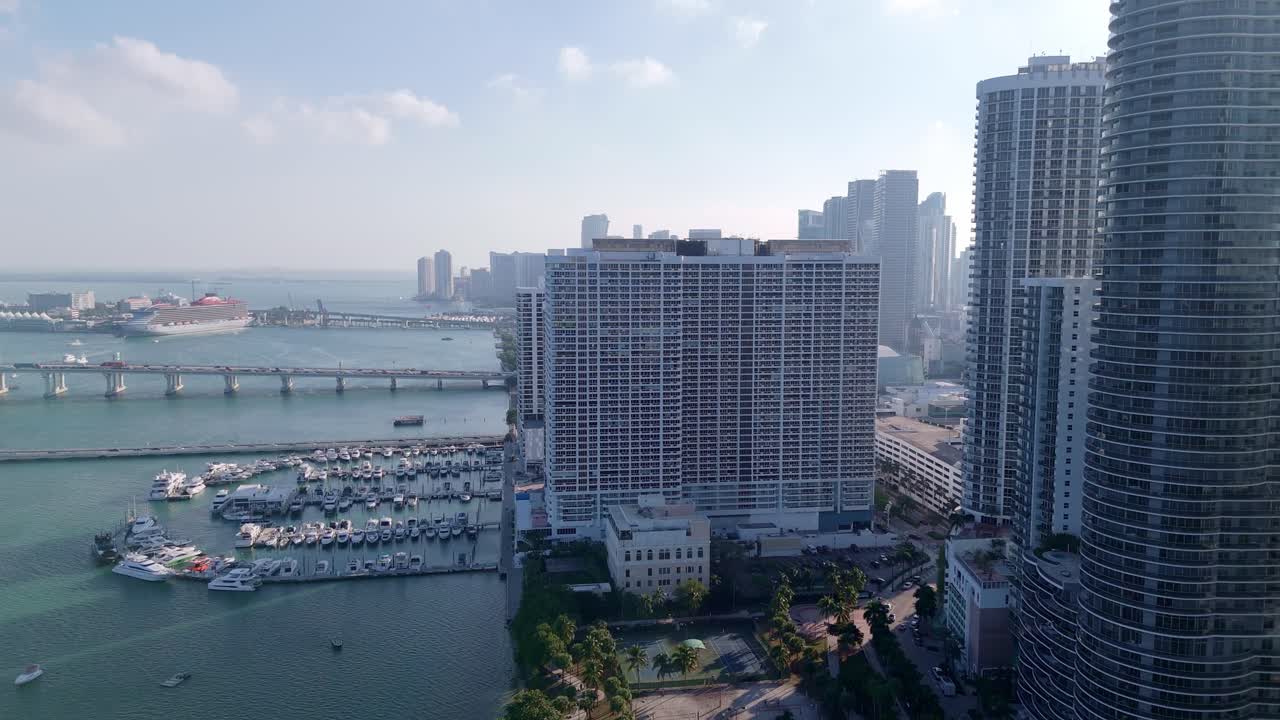 Aerial drone view of downtown Miami featuring high-rise buildings, a marina filled with luxury yachts, and Biscayne Bay with cruise ships under a partly cloudy sky.