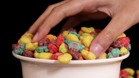 a hand reaches into a bowl of colorful cereal against a black background, highlighting vibrant colors and textures
