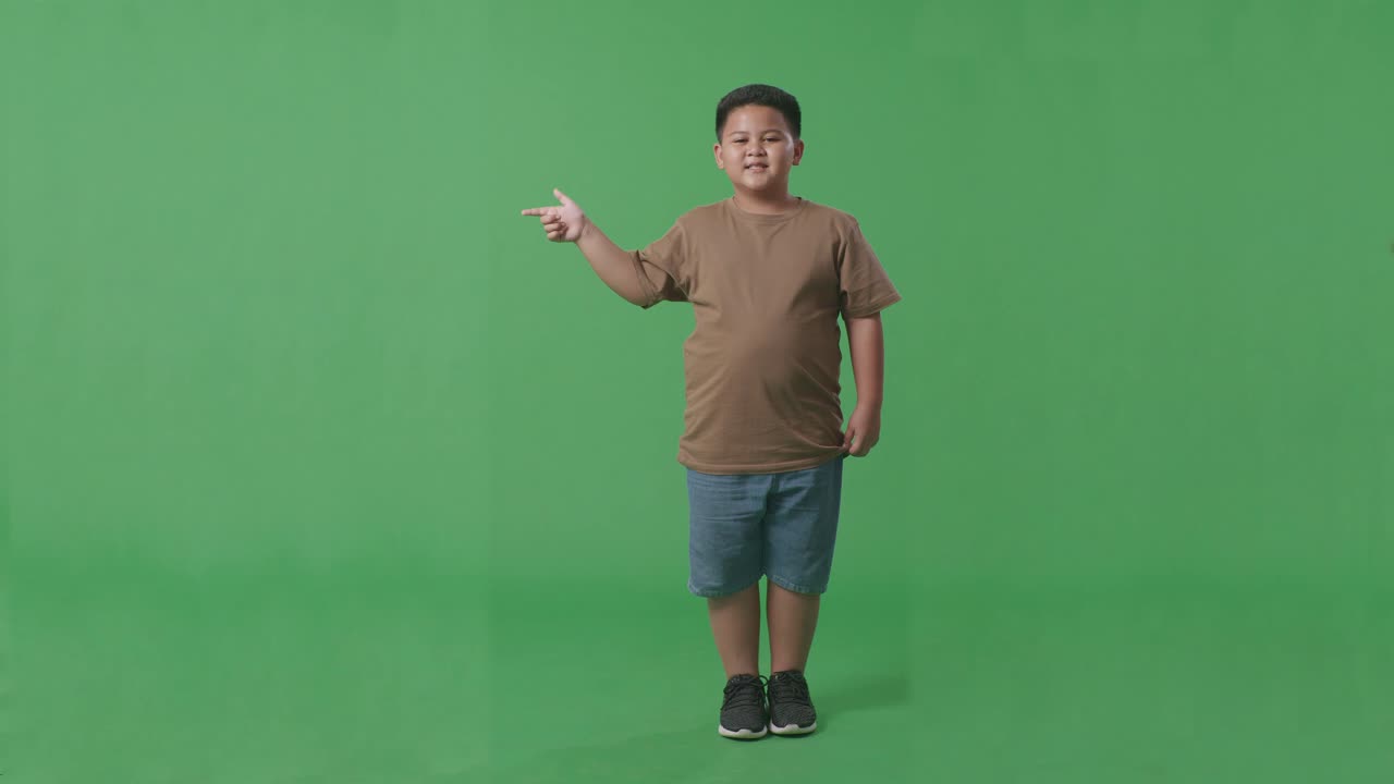 cuerpo lleno de niño asiático con una sonrisa apuntando al lado mientras está de pie en la pantalla verde