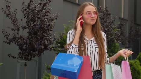 positive young girl with bags talking on mobile phone about sales in shopping mall in black friday