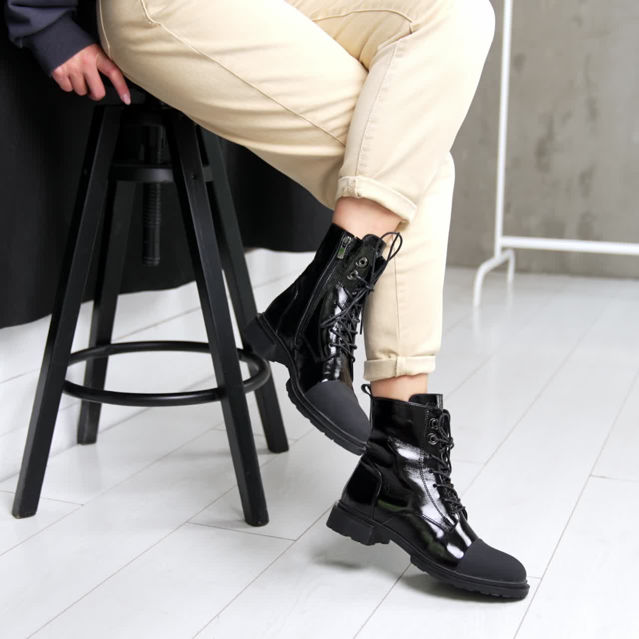 Unknown woman comes up to a high stool and sits down. Model demonstrates stylish boots from combined leather. White backdrop