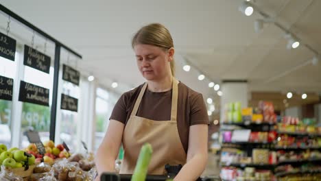 Confident-girl-in-a-brown-T-shirt-and-apron-puts-goods-on-the-counter-from-a-cart-during-her-work-in-a-supermarket