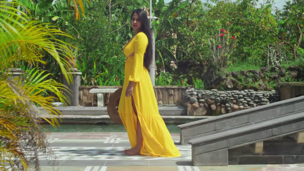A girl in a flowing yellow gown walking through the lush gardens and pool of a majestic castle