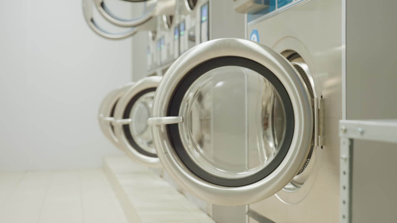 Dim laundry room with washing machines lined up, digital indicators glowing in darkness as light switches on, revealing open washer doors in industrial interior environment