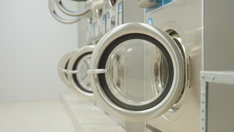 dim laundry room with washing machines lined up, digital indicators glowing in darkness as light switches on, revealing open washer doors in industrial interior environment