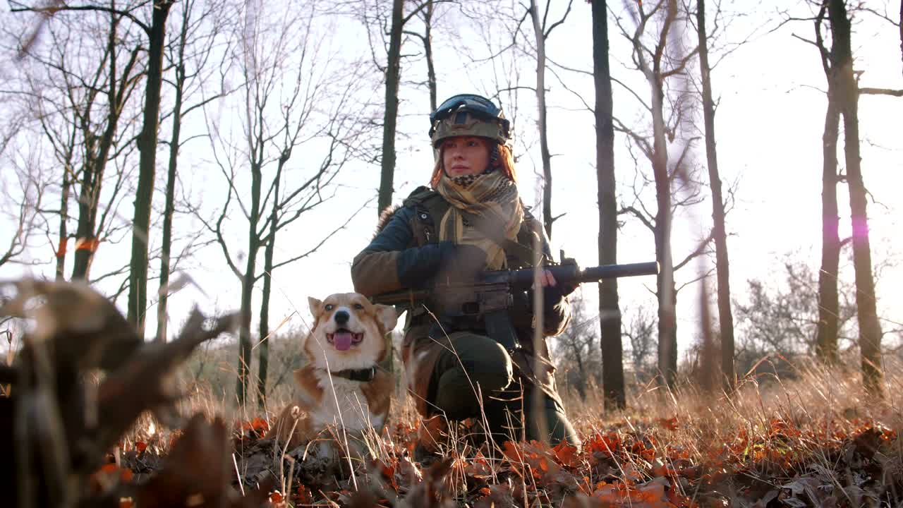 군복을 입은 젊은 예쁜 은 머리 여성은 소총으로 무장하고 개와 놀고 있습니다.
