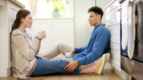 una pareja feliz hablando y bebiendo café sentada en el suelo de la cocina en casa, en cámara lenta