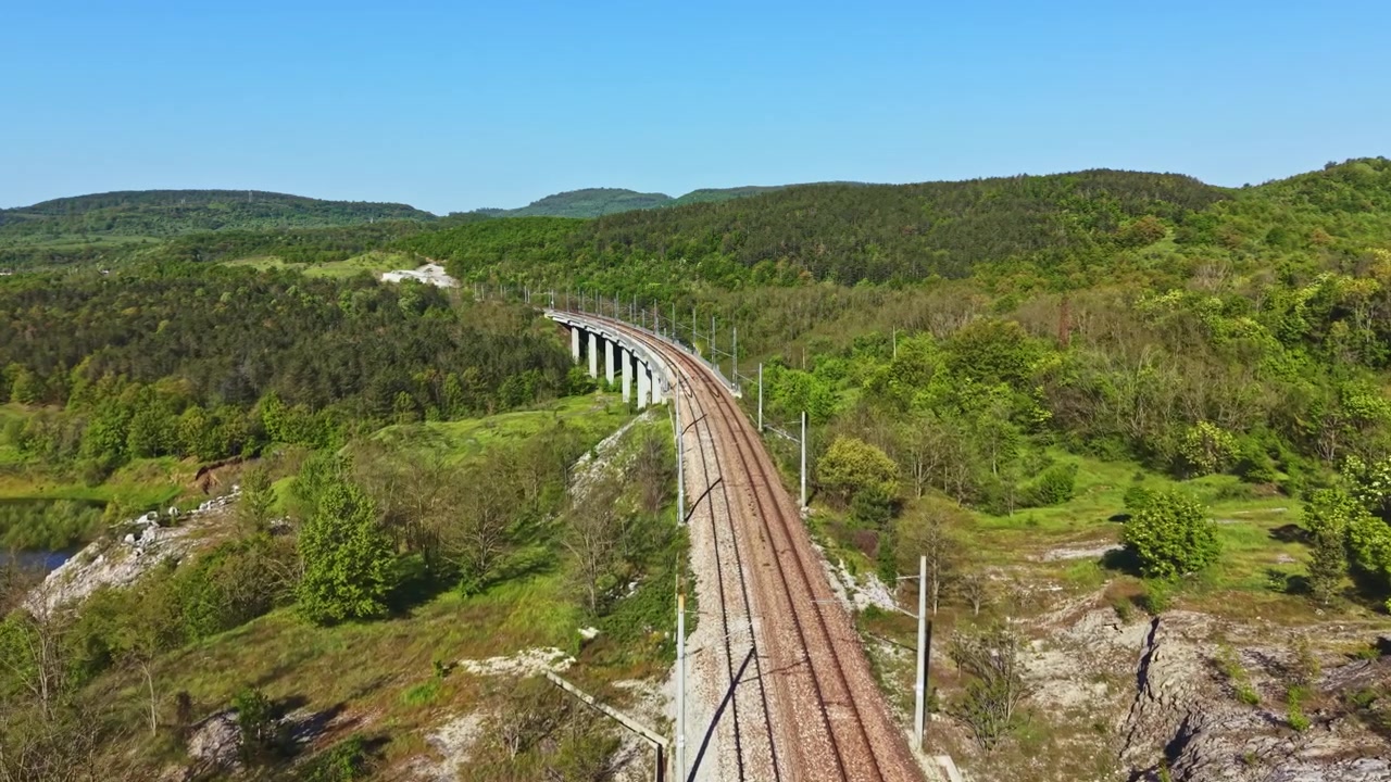 Trilhos de trem se estendem por colinas verdes perto de um rio em uma área rural em um dia ensolarado, exibindo a natureza e a engenharia humana.