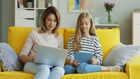 Cute-Caucasian-girl-talking-with-her-mother-while-sitting-on-the-yellow-sofa-with-laptop-computer-and-tablet-at-home.-Inside.