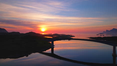 Increíble-Toma-Panorámica-Aérea-De-Un-Vehículo-Que-Pasa-Por-Los-Famosos-Puentes-Frevang-En-Lofoten,-Noruega,-Con-Aguas-Tranquilas-Que-Reflejan-La-Puesta-De-Sol-Y-Las-Montañas