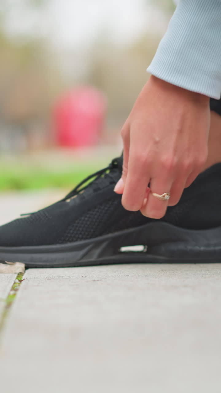 Primo piano di una signora che allaccia il laccio della scarpa destra delle sue scarpe da ginnastica nere, preparandosi per un allenamento o una corsa all'aperto, focus sulle mani che allacciano le scarpe sul marciapiede, stile di vita attivo, preparazione fisica al parco.