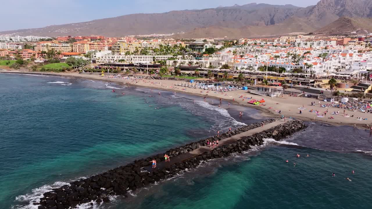 Persone nuotano e si rilassano lungo la vivace spiaggia di Fanabe con uno sfondo di città e montagne.