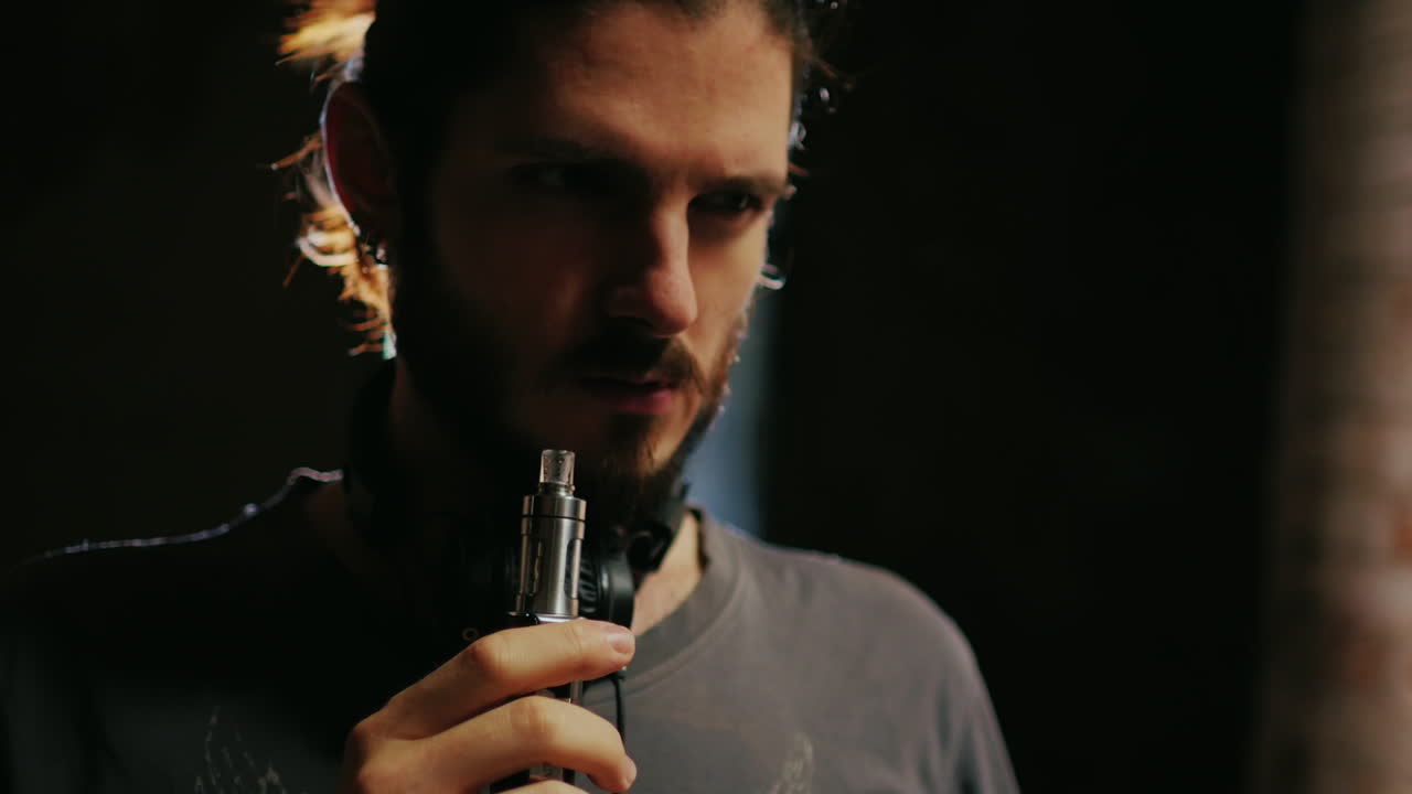un joven barbudo que fuma un cigarrillo electrónico sale maravillosamente en un haz de luz