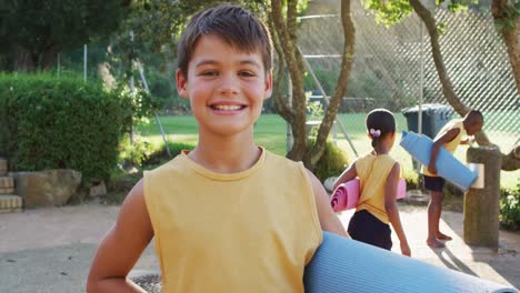 portrait of happy caucasian schoolboy holding yoga mat after yoga lesson outdoors