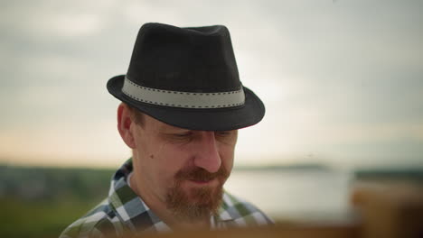 close-up of a man wearing a black hat and green checkered shirt, deeply focused on an unseen task, with mosquitoes visible around his head. the serene outdoor background adds a natural