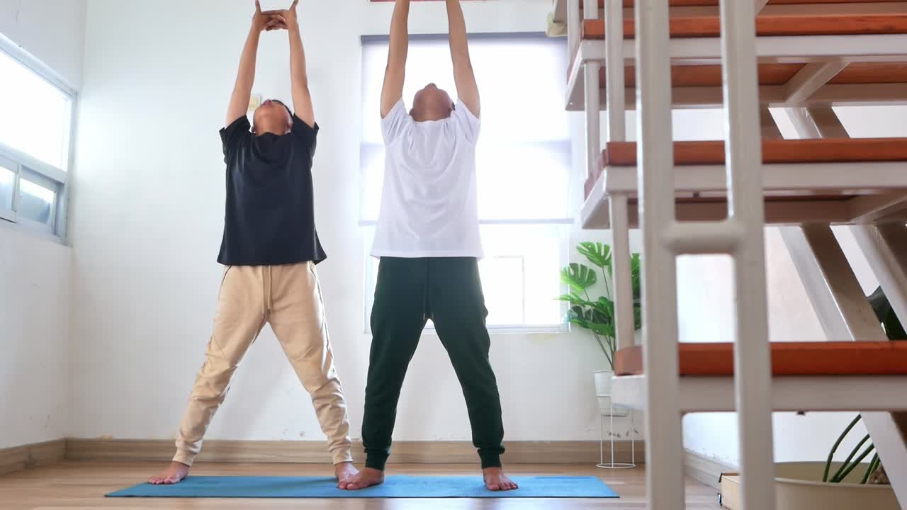 Dois irmãos adolescentes fazendo alongamentos overhead em pé e alcançando para frente em um colchonete de ioga em casa