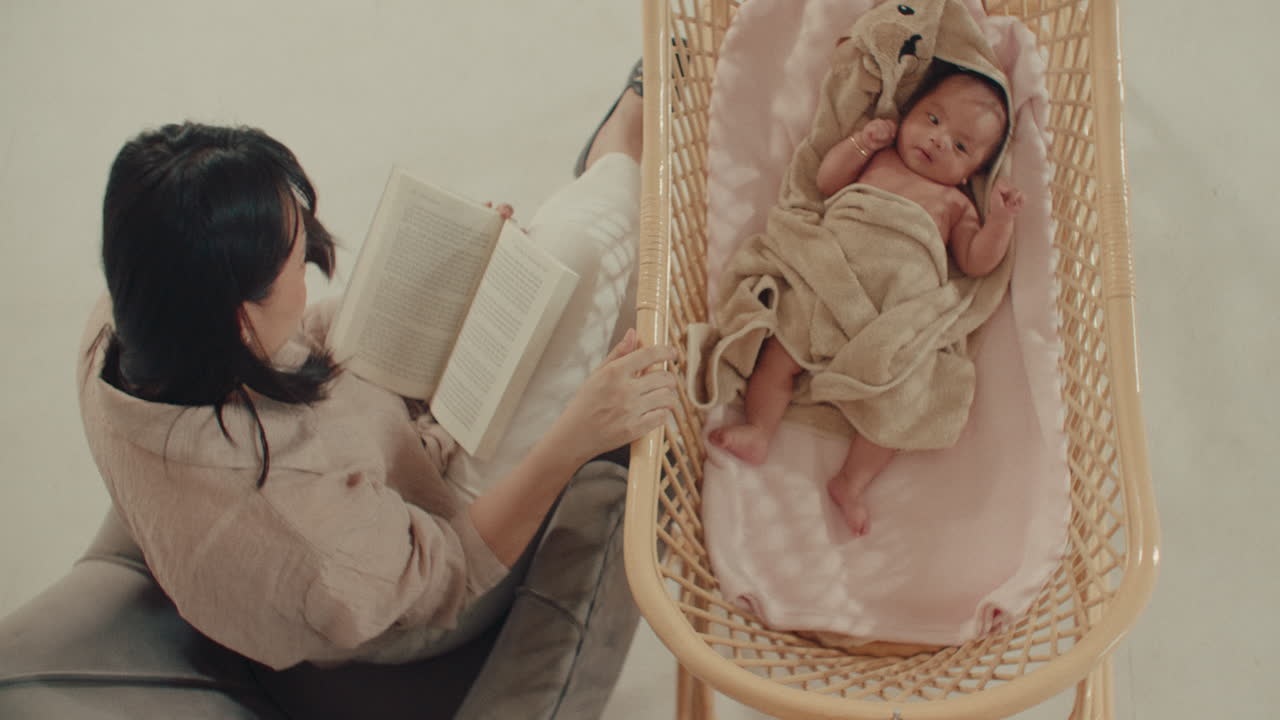 Madre che legge un libro accanto al bambino nella culla, vista dall'alto, neonato sdraiato in una culla accogliente, momento familiare sereno