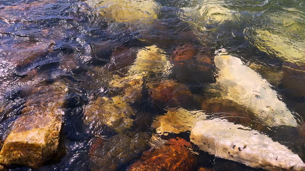 A Serene Underwater Scene Captured: Clear Waters Revealing a Bed of Colorful Stones Below, Illuminated by Natural Light and Gentle Ripples Across the Surface
