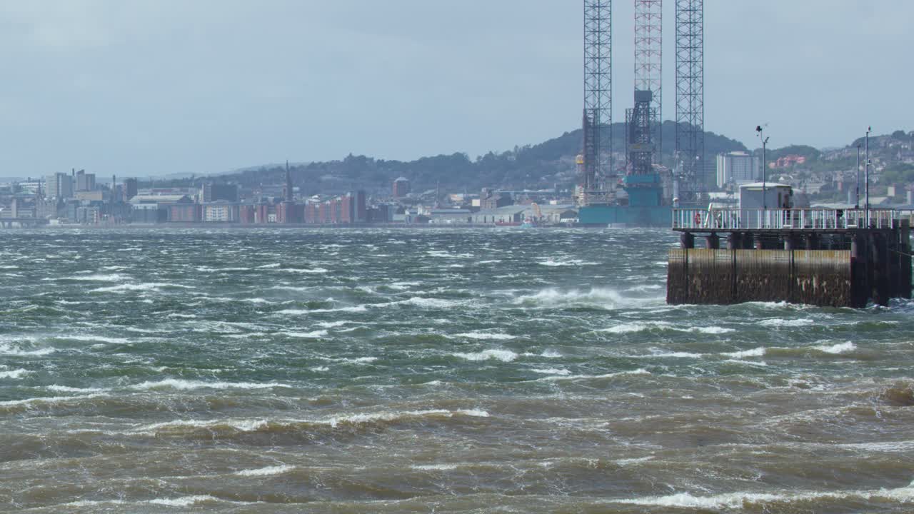Choppy sea waves hit concrete pier under cloudy daylight, distant cityscape and offshore platform visible