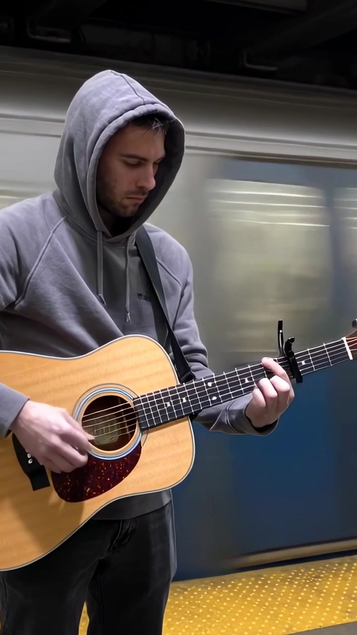 Video verticale: Uomo con felpa grigia che suona la chitarra con un capotasto su una banchina della metropolitana, con un treno che passa