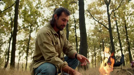 a brunette man in a light green jacket puts firewood in a small fire during a halt on a hike in a yellow-green autumn forest with withered grass. happy man resting during a rest near a fire while hiking in the forest