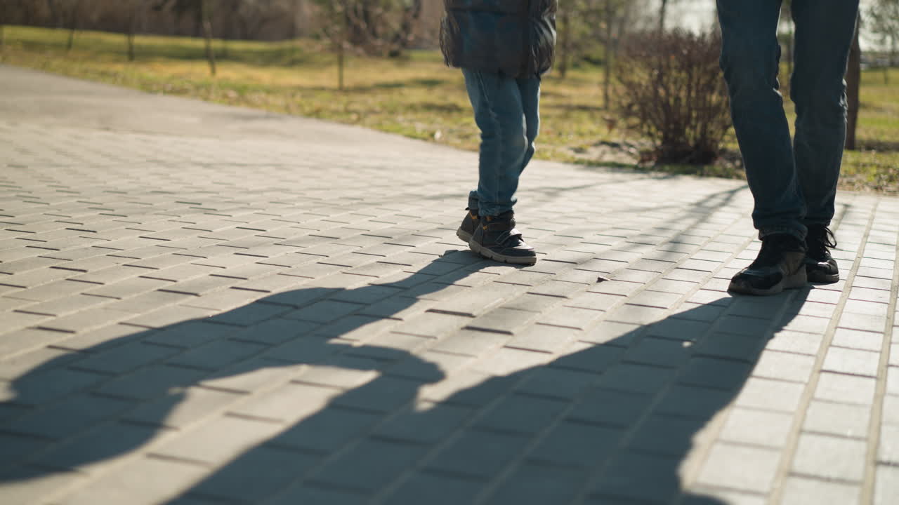 A close-up of two people, an adult and a child, playfully moving their legs on a paved path, both are wearing blue jeans and boots, their shadows stretching across the pavement