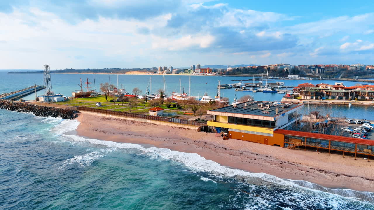 Luftpanorama af sandstrand og havn med yachter. Bred droneudsigt over kystlinjen med bølgebryder, strand og lystbådehavn mod overskyet himmel.