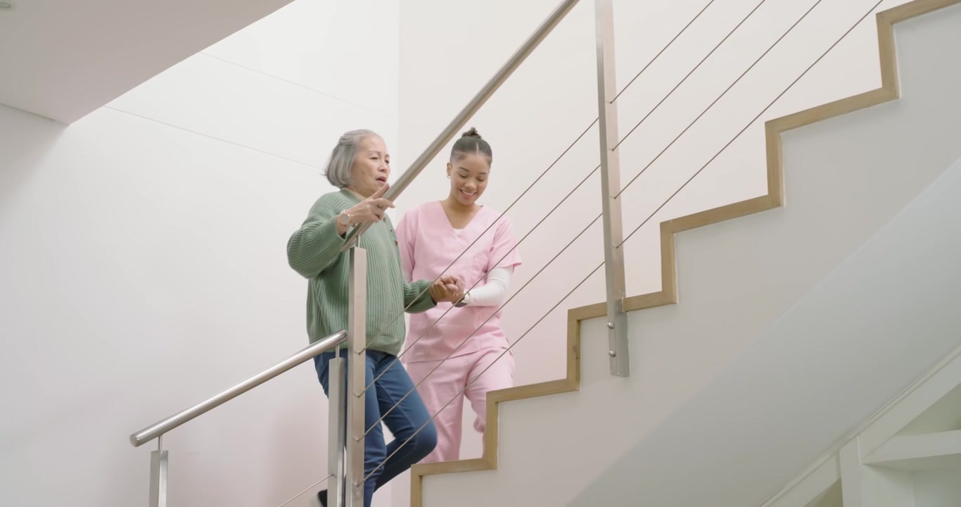 Assistente in divisa rosa che prende il braccio di una donna anziana, guidandola su per le scale di legno usando il corrimano per l'equilibrio.