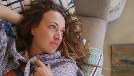 caucasian woman lying on sofa with blanket, relaxing and looking away in cottage living room