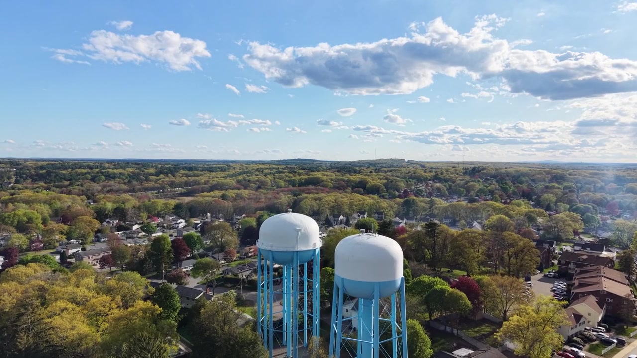 Two blue water towers rising above dense autumn tree canopy, suburban neighborhood and busy road visible through fall foliage under blue sky with white clouds. Massachusetts, USA. Aerial approachingx