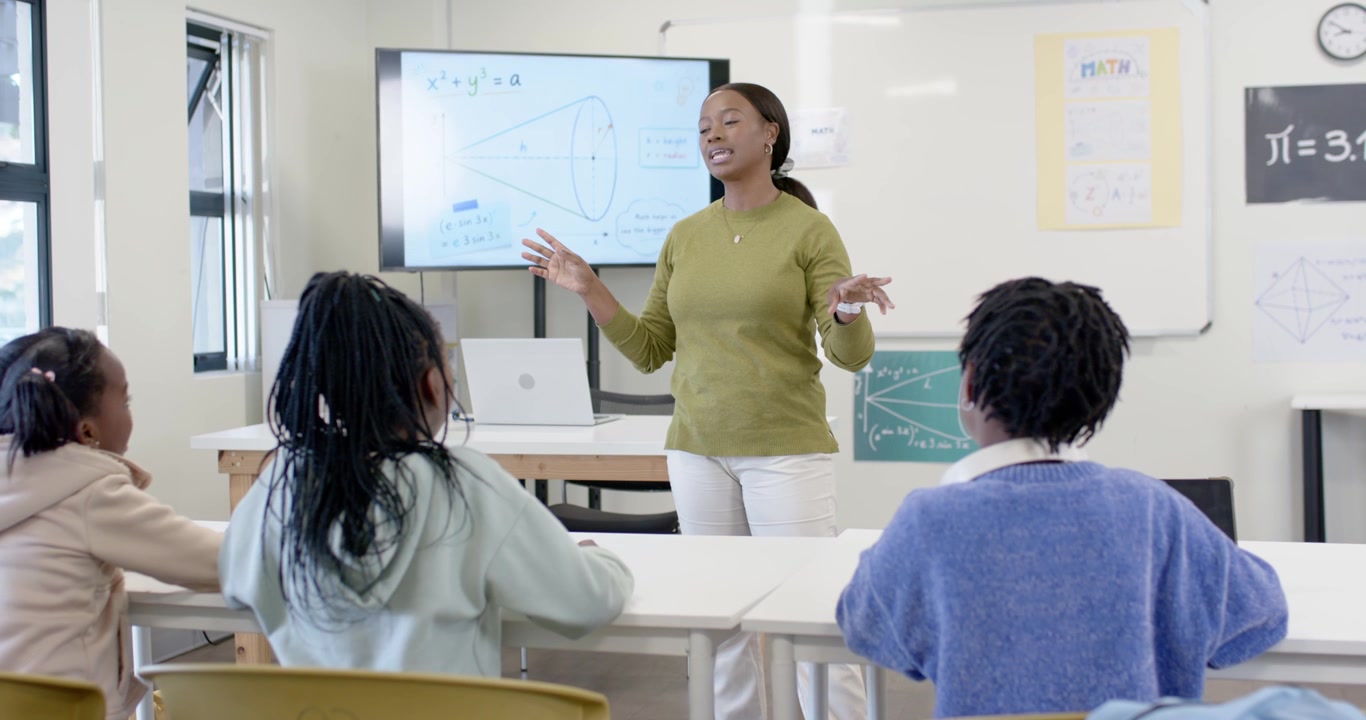 African woman teacher pointing at display, showing math and teaching three African girls in class