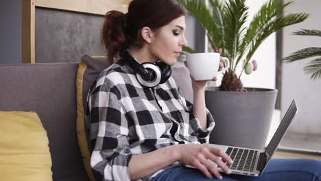 Relaxed,-charming-girl-sits-on-a-sofa-in-the-room,-types-on-a-laptop.-A-big-white-cup-in-her-hand.-The-girl-drinks-cappuccino-and-enjoys-the-time.-Headphones-on-the-neck