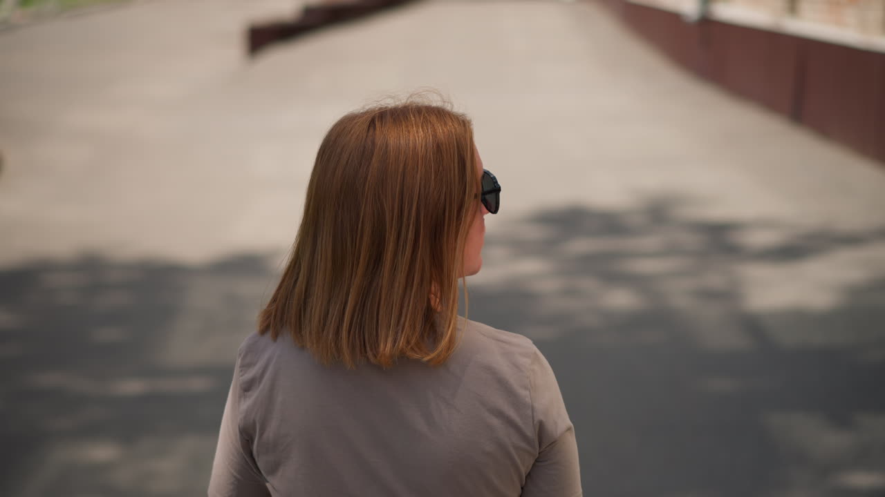 Behind view of student with golden hair wearing sunglasses walking fast along sunny street checking time on smartwatch while looking around, soft light and blurred shadows
