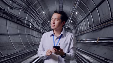 asian male professional worker standing with his smartphone in underground subway tunnel, he observes by looking around before he come to concentrating with his smartphone