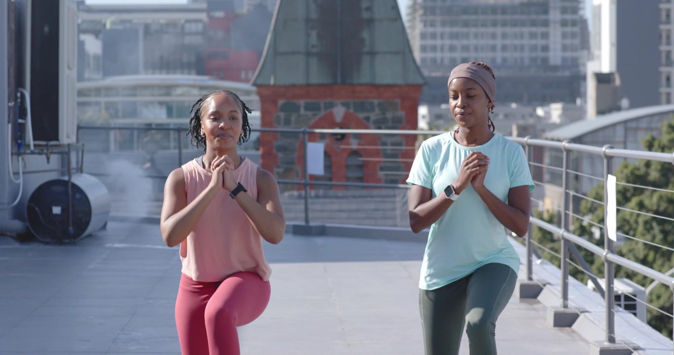 Donne afroamericane che fanno sollevamenti delle gambe in sincronia con le mani giunte su un tetto, indossando orologi per il fitness.