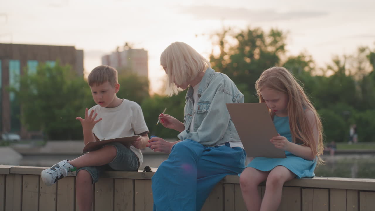 elegant mom seated on wooden dock between toddlers gently takes brush from boy to dip into paint jar while girl intently paints on board with pool and trees glowing under soft sunset sky
