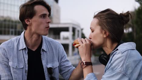 Hermosa-Pareja-Joven-Disfrutando-De-Una-Comida-Juntos.-Comiendo-Hamburguesas-Juntas-Por-Los-Lados.-Anochecer,-Lado-Del-Parque