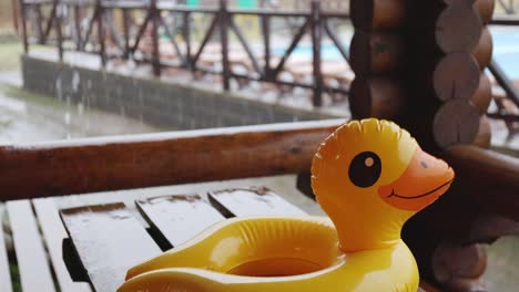 an inflatable duck lies on wooden table in gazebo at recreation center against the backdrop of pool covered with rain