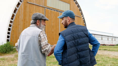 Rear-view-of-old-father-and-son-farmers-walking-and-talking-outside-the-stable