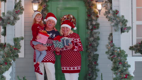 Felices-Abuelos-Mayores-Con-Nieta-Sosteniendo-Muchas-Cajas-De-Regalo-Cerca-De-La-Casa-De-Navidad,-Celebran