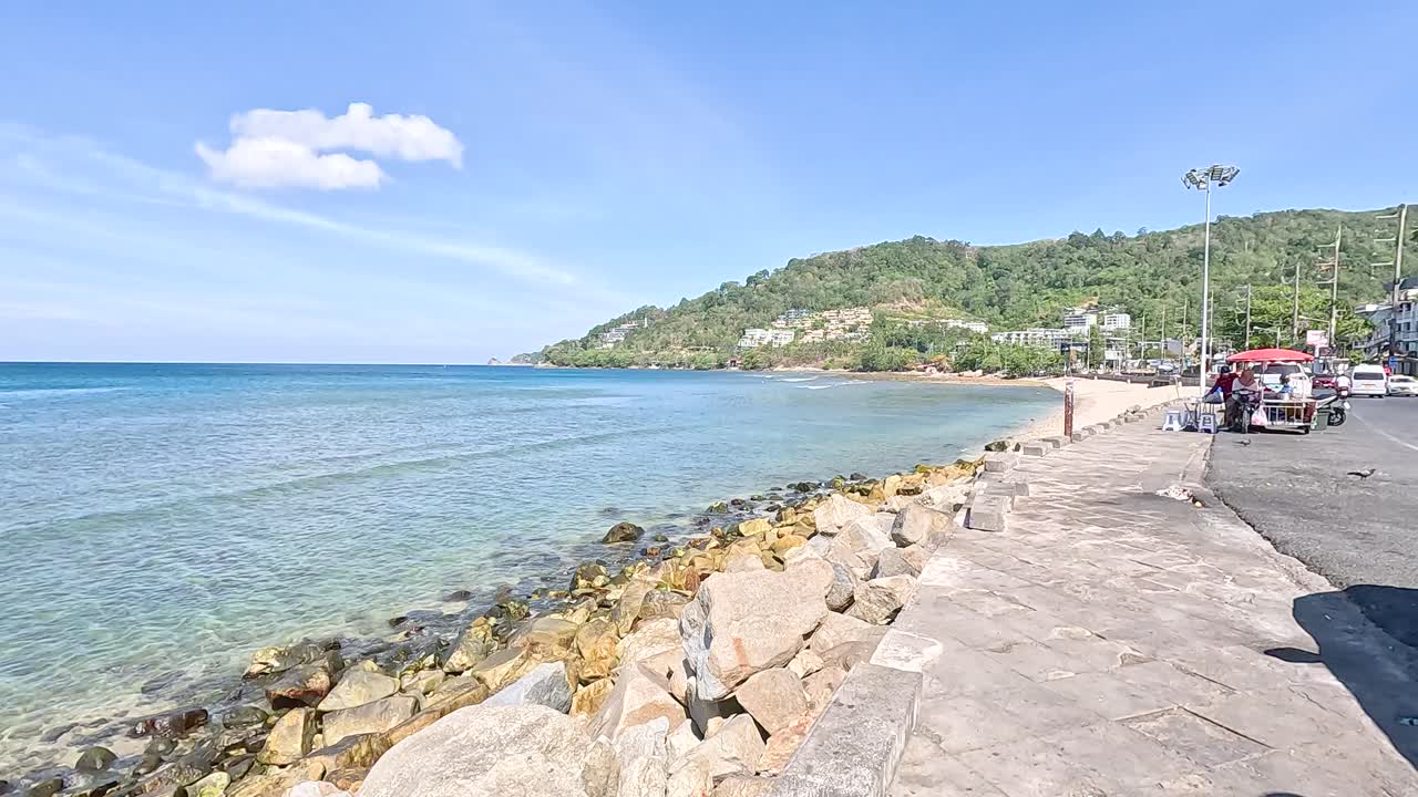 A serene coastal scene at Kalim Beach, Phuket, with clear skies, calm waters, and a rocky shoreline under bright sunlight