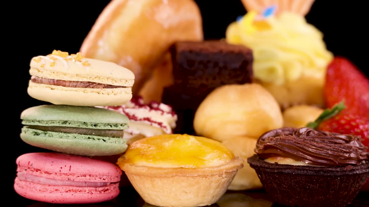 Hand selects a colorful macaron from assorted bakery desserts under bright studio lighting, black background