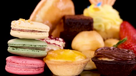 hand selects a colorful macaron from assorted bakery desserts under bright studio lighting, black background